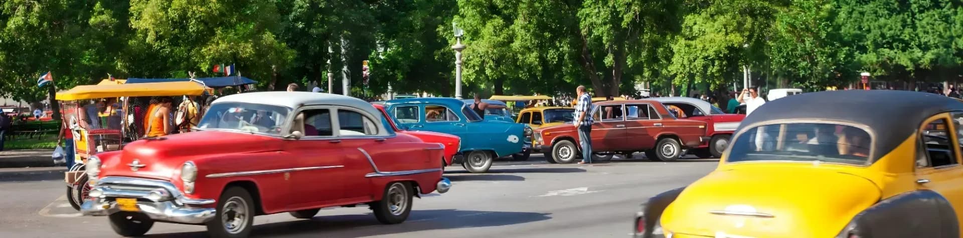 Vintage cars line a street in a leafy, urban setting. A Cuban flag flies in the background.