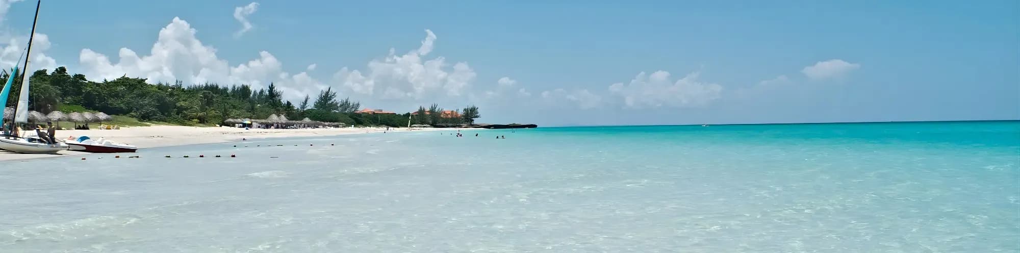 Turquoise waters meet a white sand beach lined with palm trees and thatched umbrellas. Sailboats and small boats are pulled ashore.