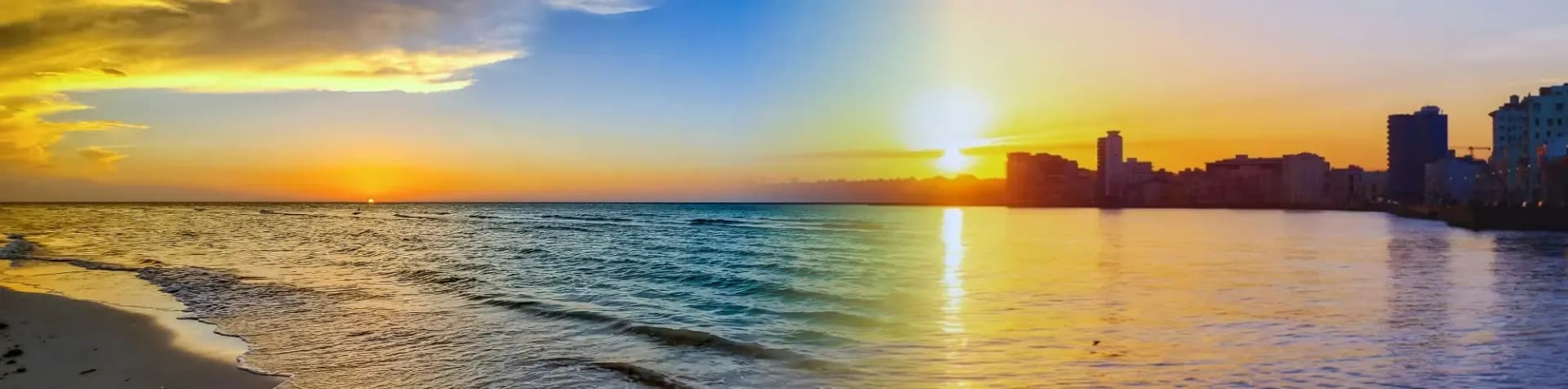 A panoramic view of a sunset over a beach and a city skyline reflected in the water.