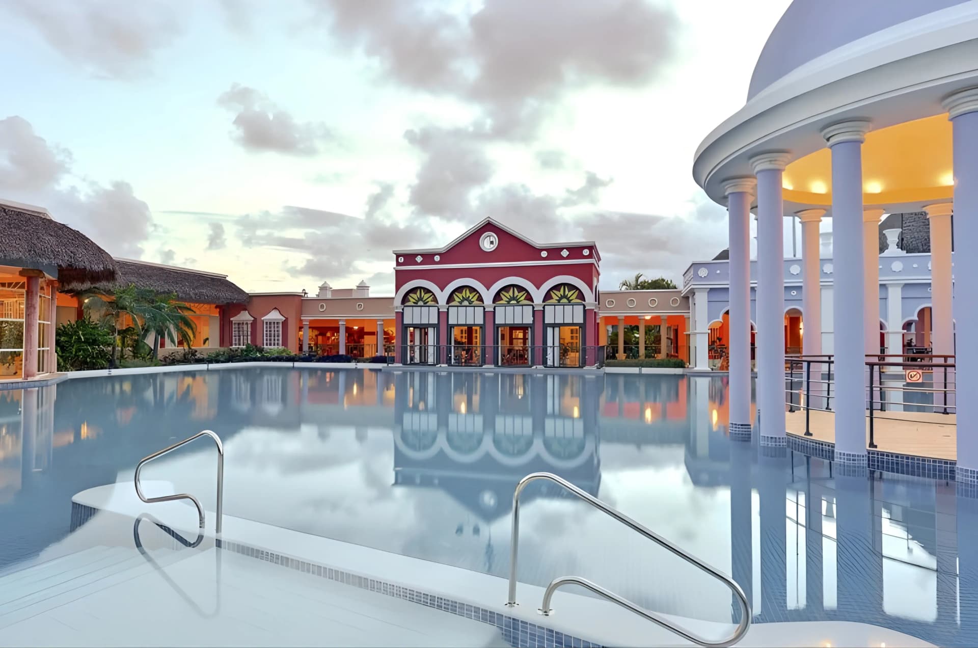 A serene resort pool reflects the pastel-colored buildings and cloudy sky at dusk, with a thatched-roof structure on the left and a domed building with columns on the right.