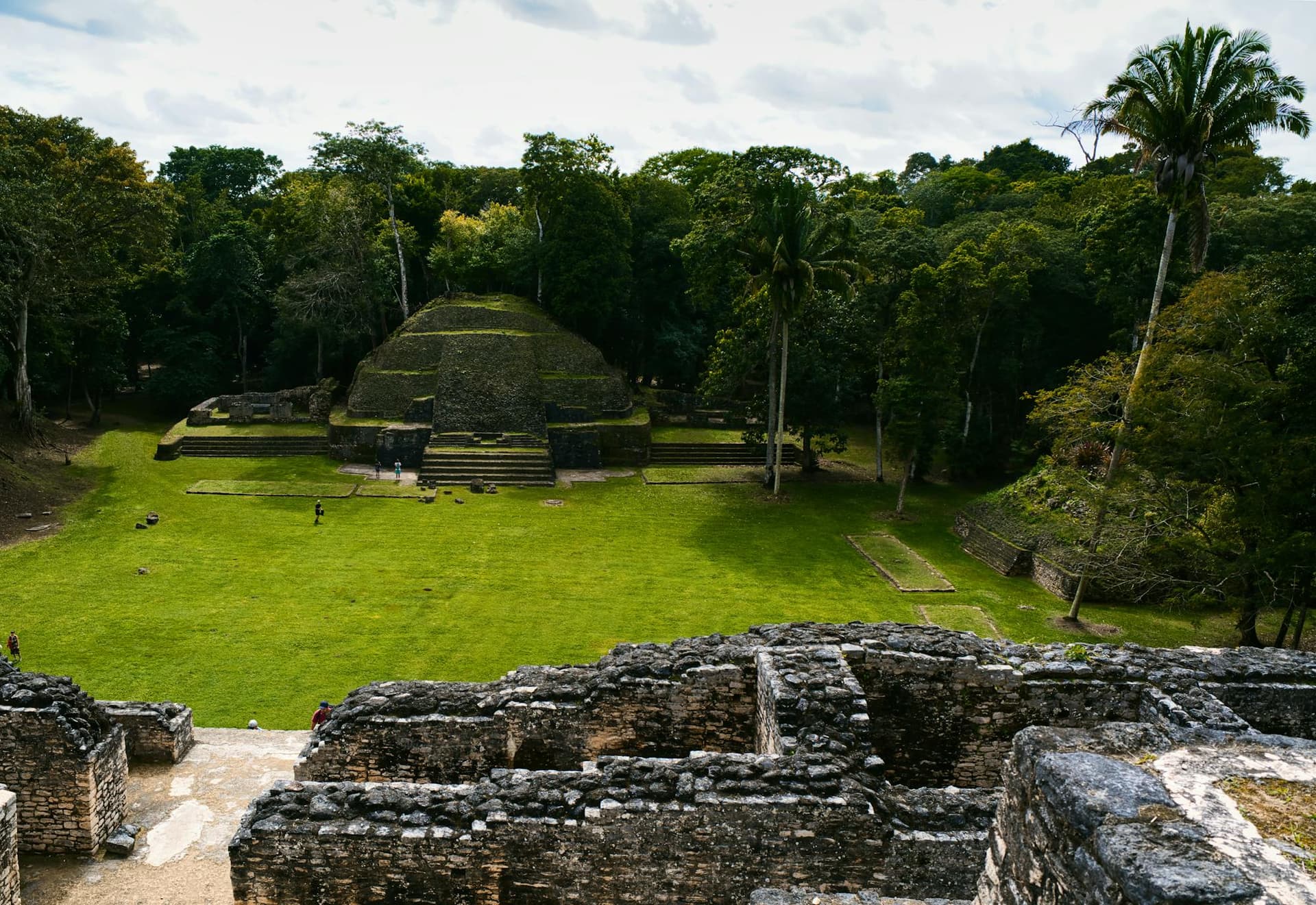 A stepped ancient Maya pyramid rising from a green plaza ringed by dense jungle and palms in Belize