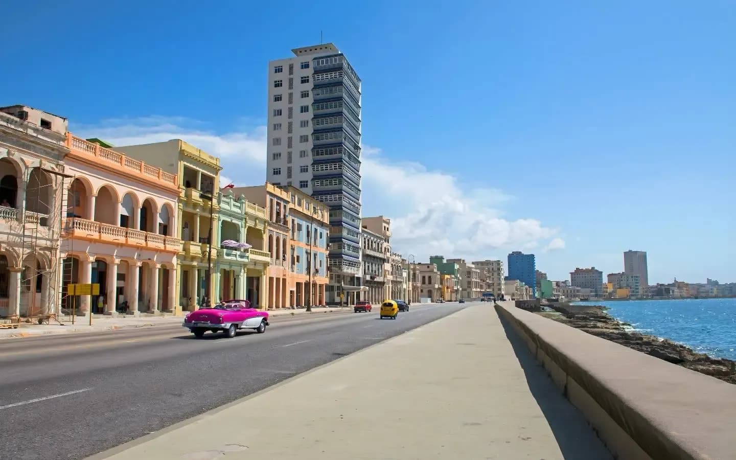 A vintage pink convertible drives along a coastal road lined with colorful colonial buildings under a bright blue sky.
