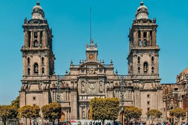 The Metropolitan Cathedral of Mexico City stands majestically under a clear blue sky, its twin bell towers flanking a central facade adorned with intricate carvings and a prominent clock.