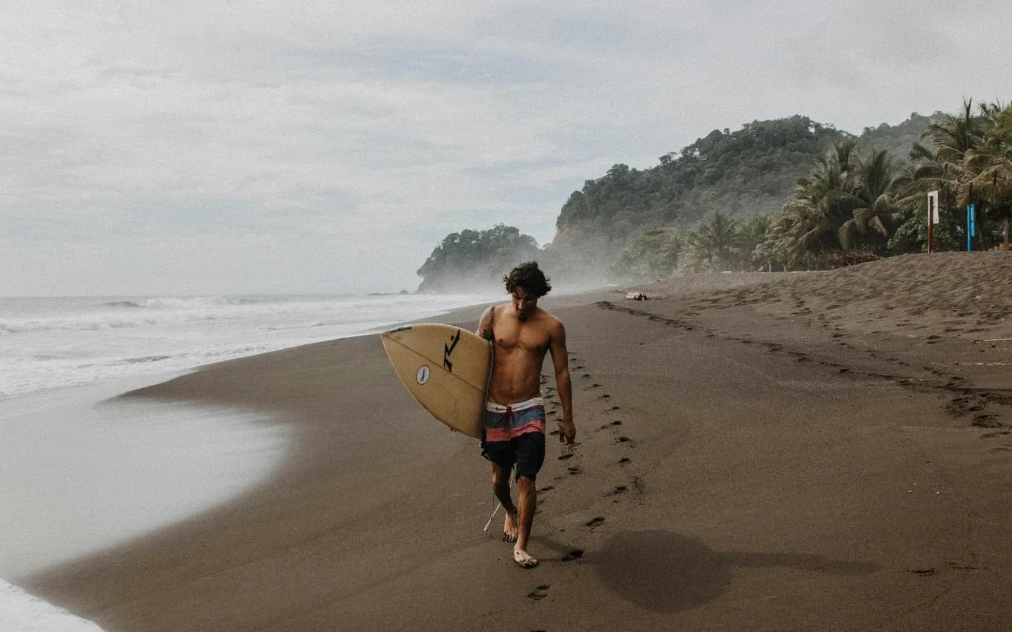 Surfer walking along a dark-sand Pacific beach in Costa Rica
