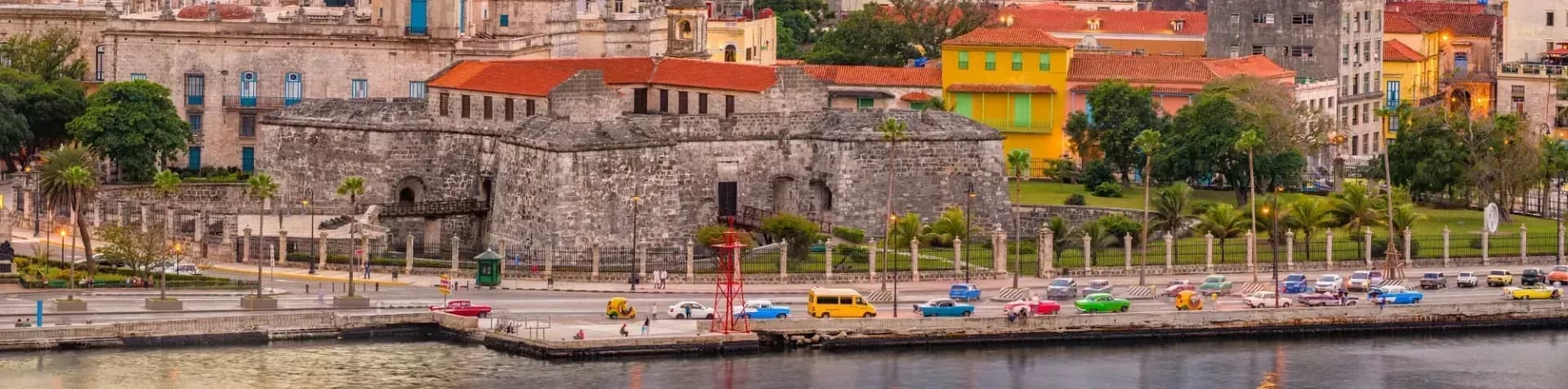 Historic stone fortress and colorful buildings line a waterfront street with vintage cars in Havana, Cuba.