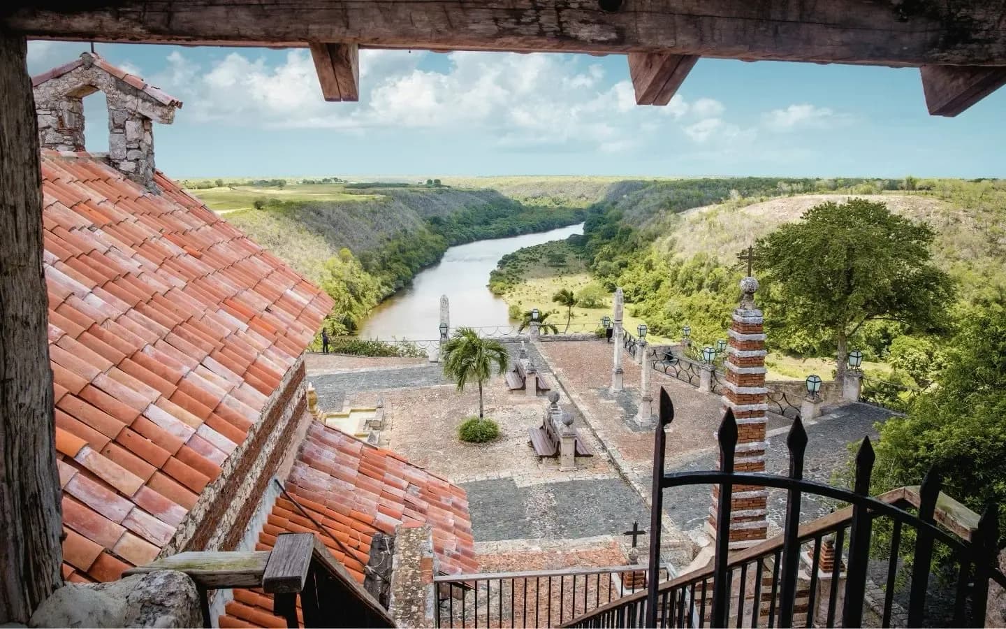 A view from a wooden balcony overlooking a red-tiled roof, a courtyard with benches, and a winding river through a lush green valley under a cloudy sky.