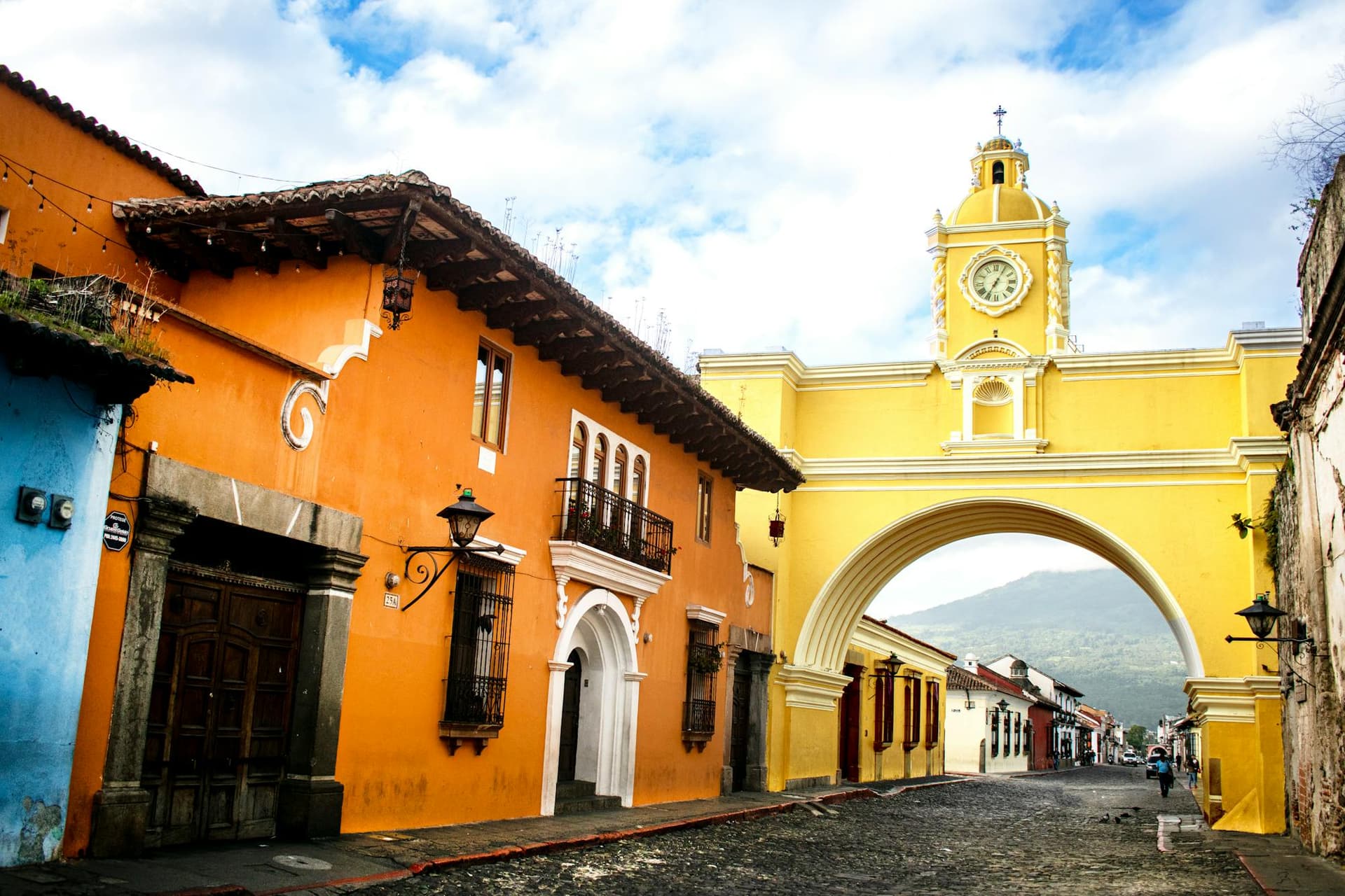 The yellow Arco de Santa Catalina and clock tower on a cobbled colonial street in Antigua Guatemala, with a volcano framed beyond