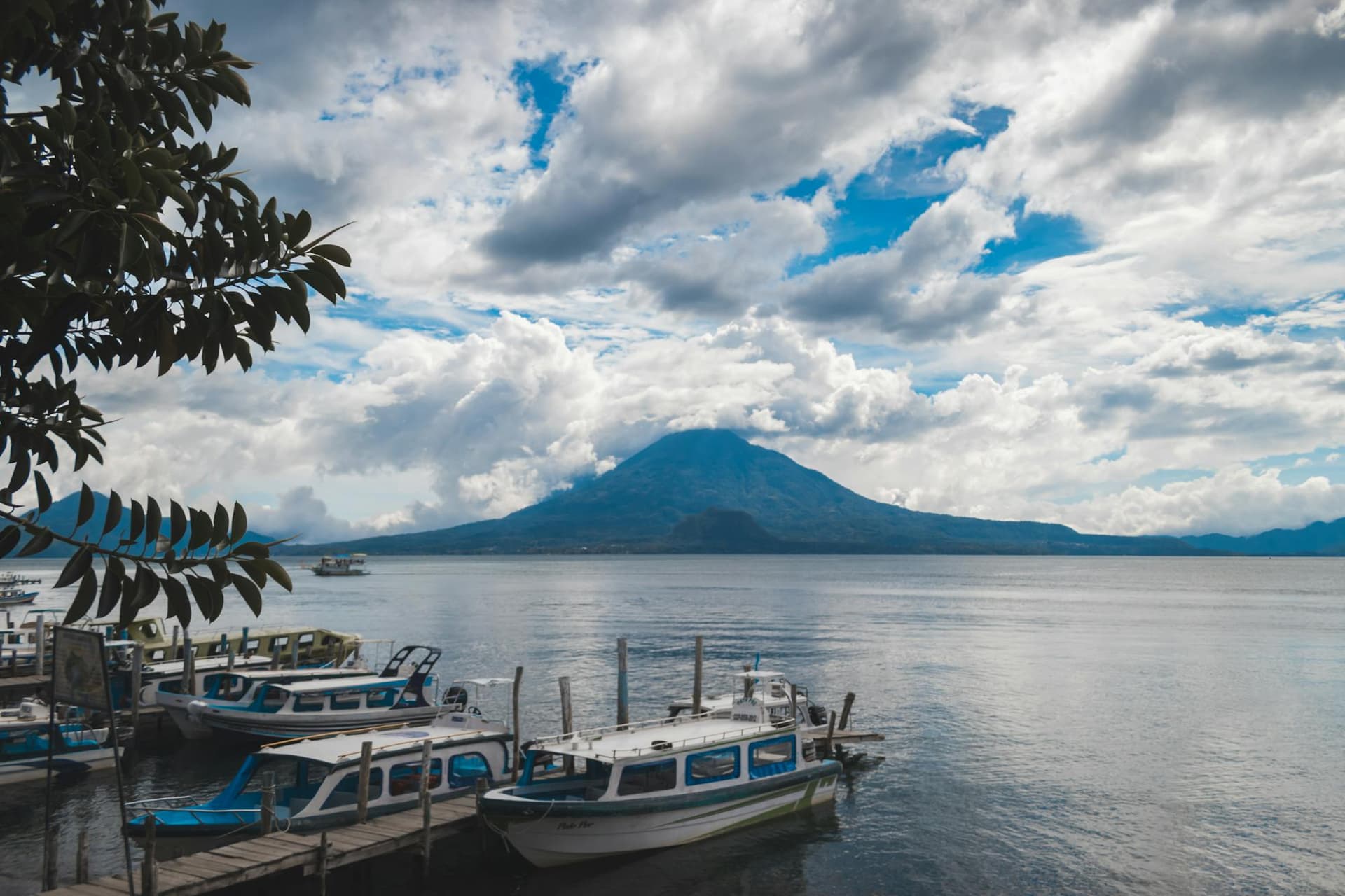 Blue and white boats moored on Lake Atitlán with a volcano rising under dramatic clouds, Guatemala