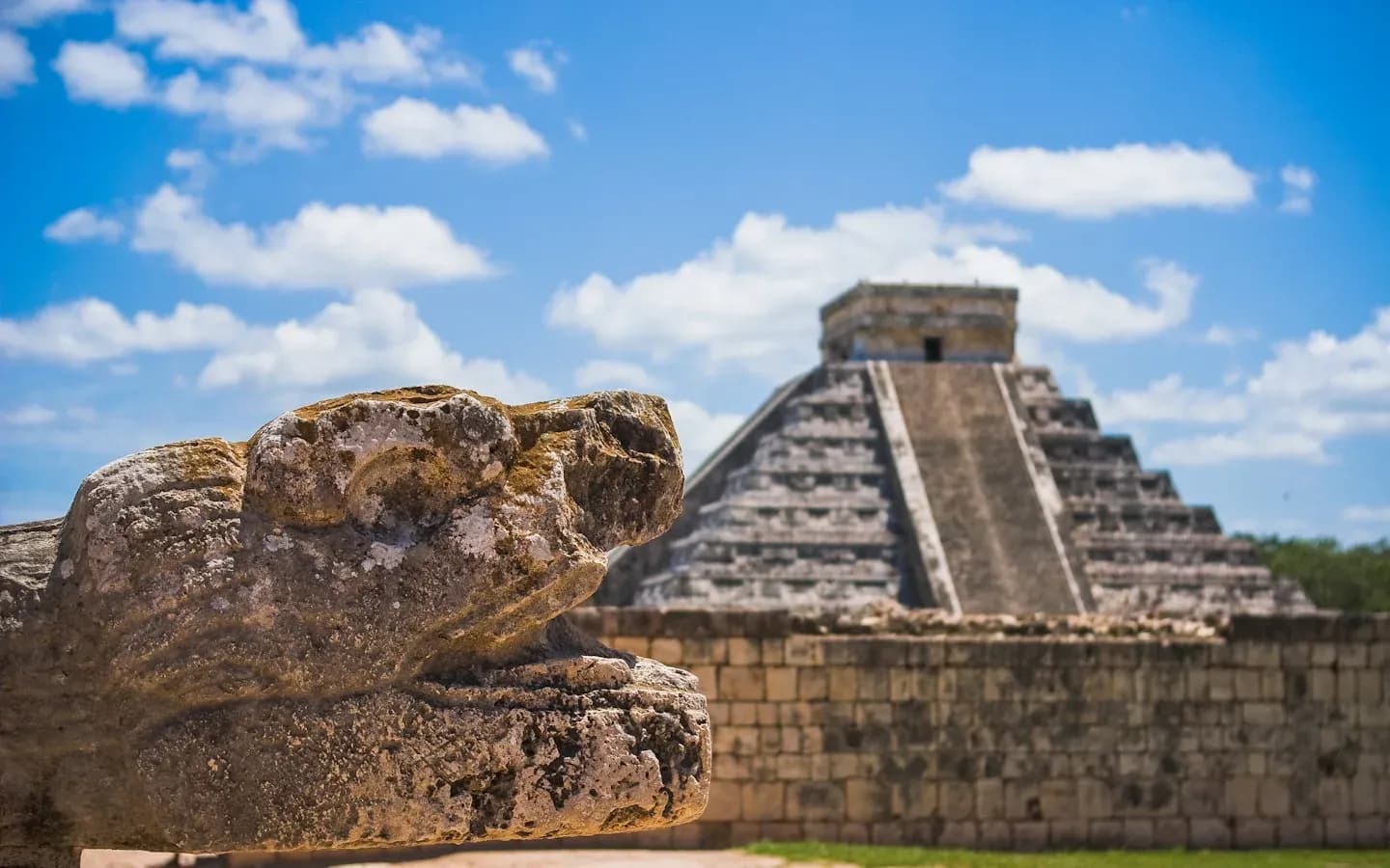 A stone serpent head sculpture in the foreground with the El Castillo pyramid at Chichen Itza in the background under a blue, cloudy sky.