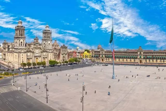 A panoramic view of the Zócalo in Mexico City, featuring the Metropolitan Cathedral and the National Palace under a bright blue sky with scattered clouds.