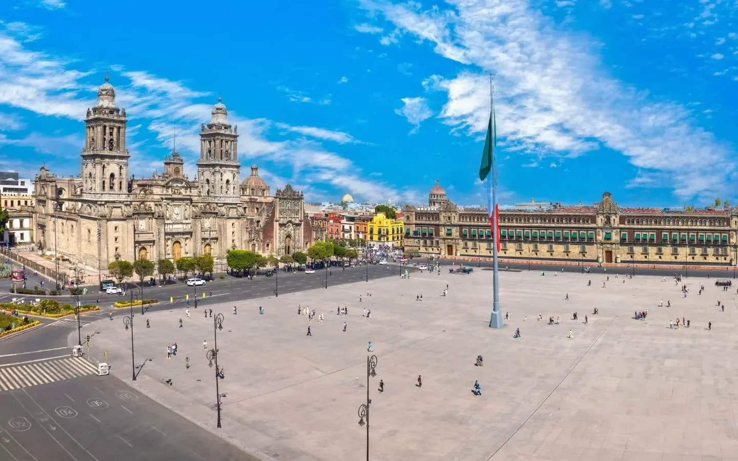 A panoramic view of the Zócalo in Mexico City, featuring the Metropolitan Cathedral and the National Palace under a bright blue sky with scattered clouds.