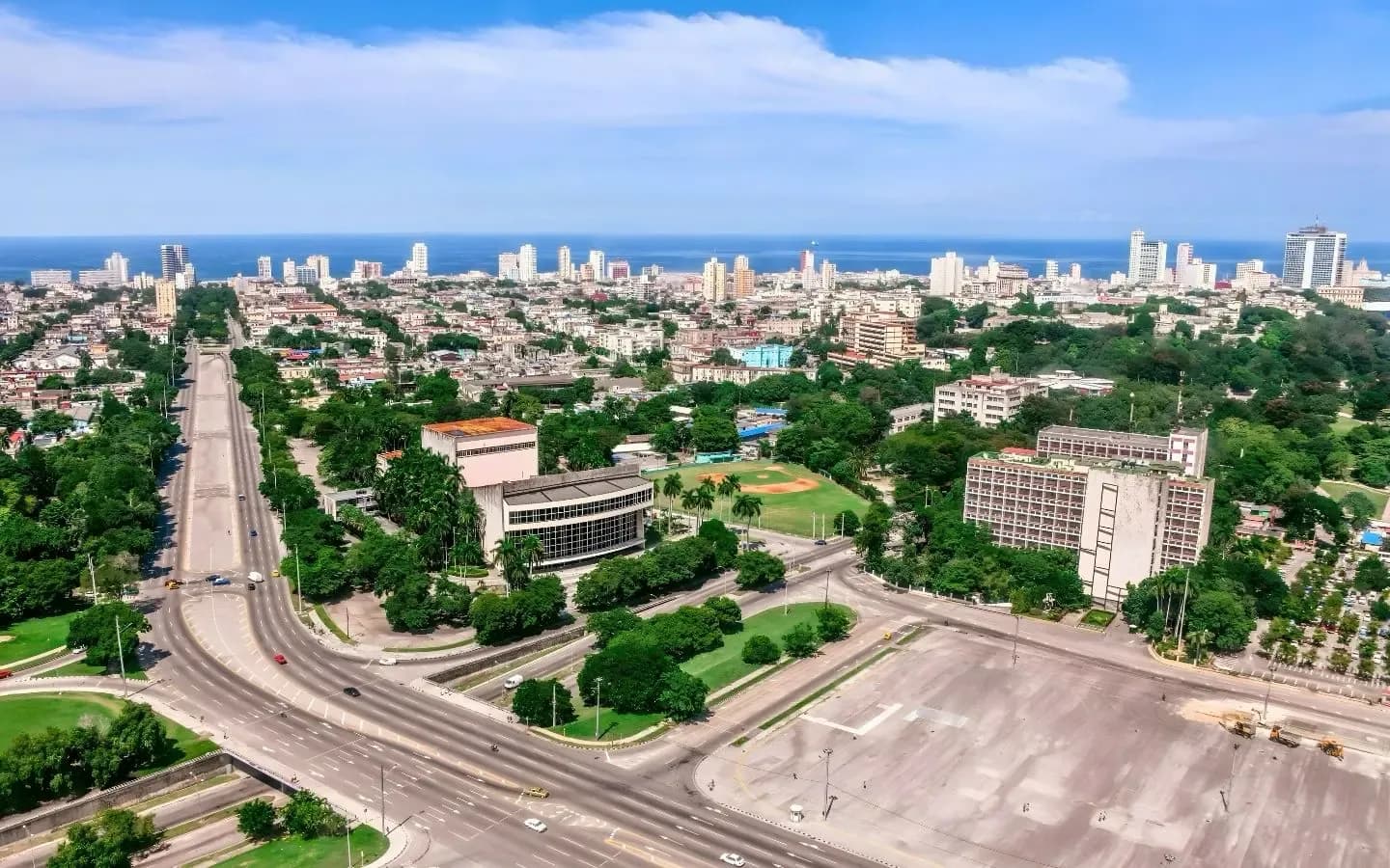 Aerial view of a wide, tree-lined avenue leading to a coastal city skyline with modern buildings and the ocean in the background.