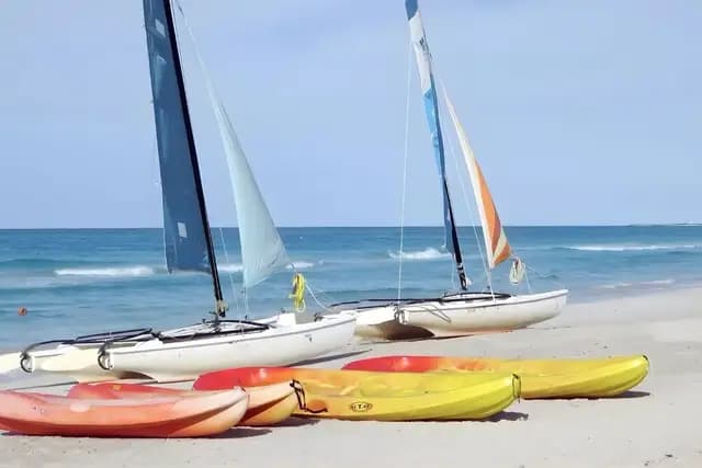 Two white catamarans with sails are parked on a sandy beach, with three colorful kayaks in the foreground and the blue ocean and sky in the background.