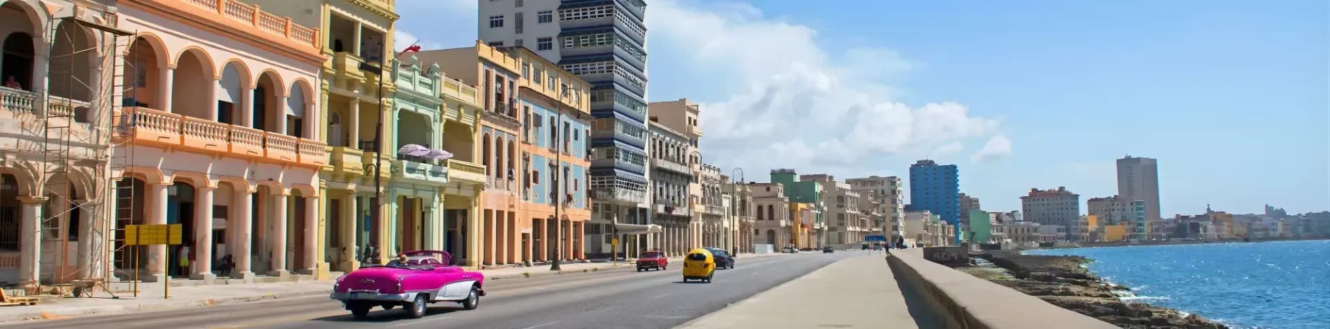 Colorful vintage cars drive along a seaside promenade lined with historic buildings.