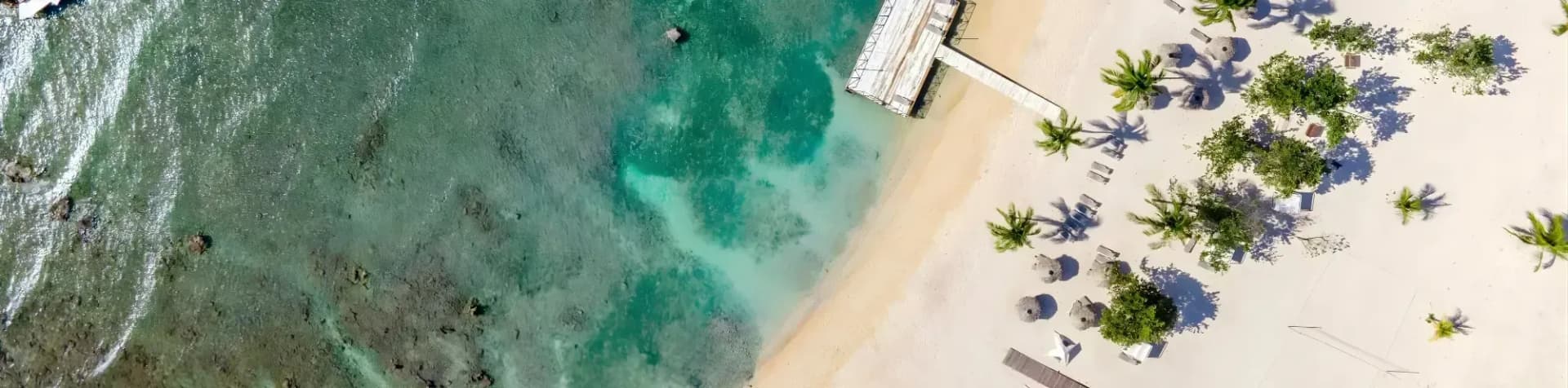 Aerial view of a tropical beach with clear turquoise water, a wooden pier, and palm trees.
