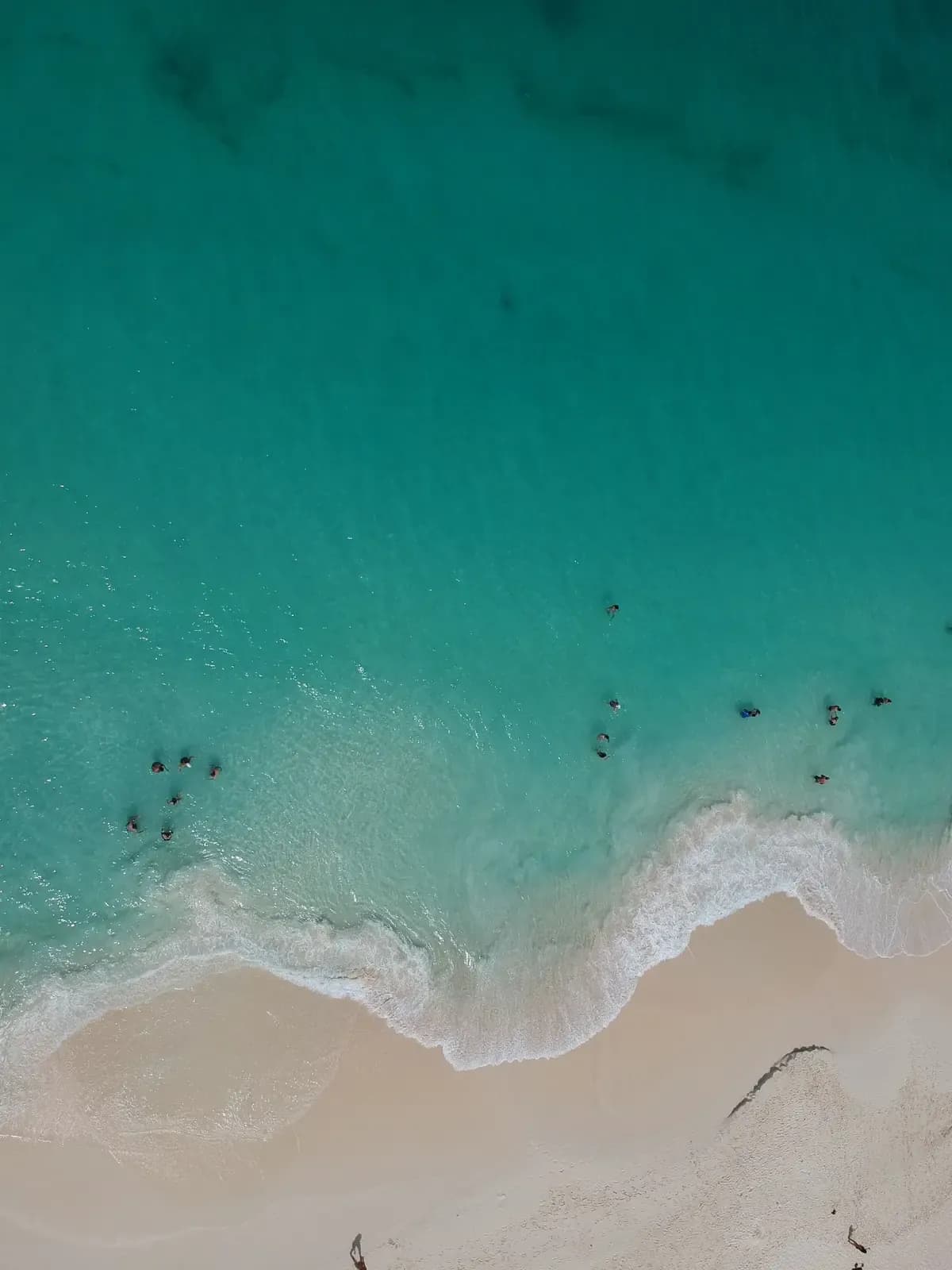 Looking straight down on clear turquoise water breaking onto the white sand of Eagle Beach, Aruba, with a few swimmers in the shallows