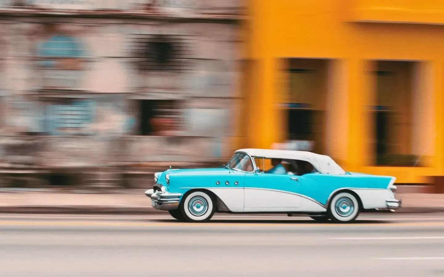 A vintage turquoise and white convertible classic car speeds down a street with motion blur, passing a blurred yellow building.