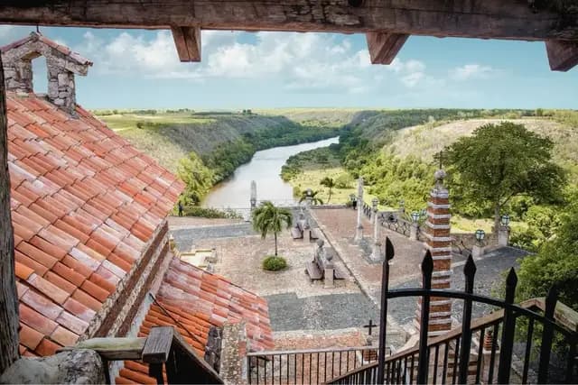 A view from a wooden balcony overlooking a red-tiled roof, a courtyard with benches, and a winding river through a lush green valley under a cloudy sky.