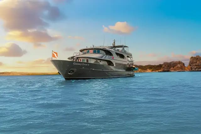 Galaxy Diver II at anchor at sunset off a Galápagos coastline, hull name visible on the bow