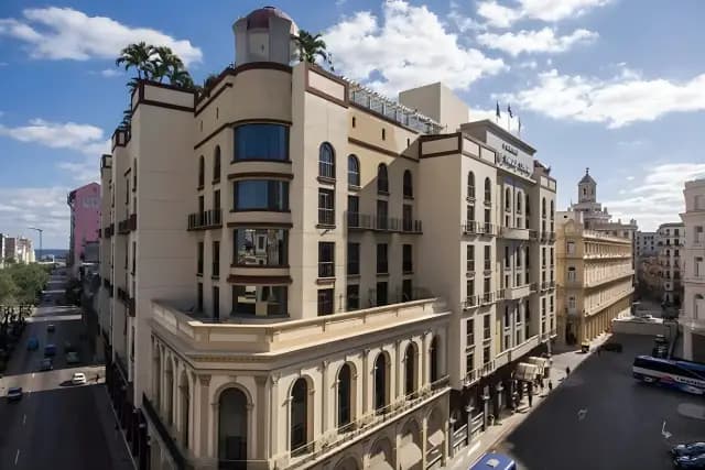 An aerial view of a cream-colored hotel with a domed tower, palm trees on its roof, and arched windows, situated on a street with cars and a blue bus in a sunny, cloudy urban environment.