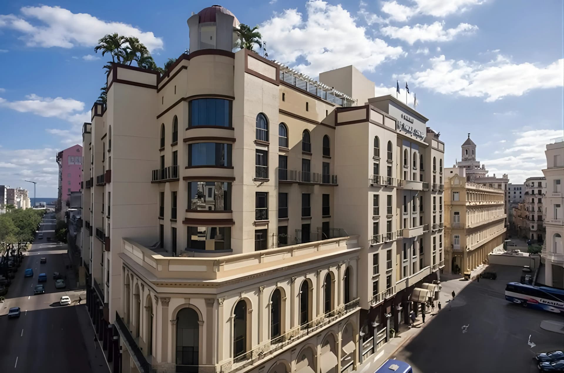 An aerial view of a cream-colored hotel with a domed tower, palm trees on its roof, and arched windows, situated on a street with cars and a blue bus in a sunny, cloudy urban environment.