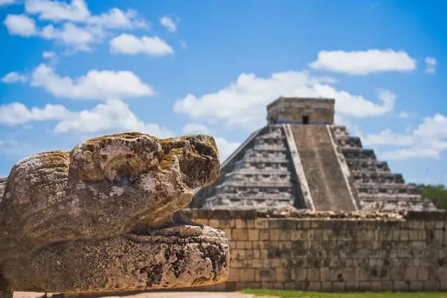 A stone serpent head sculpture in the foreground with the El Castillo pyramid at Chichen Itza in the background under a blue, cloudy sky.
