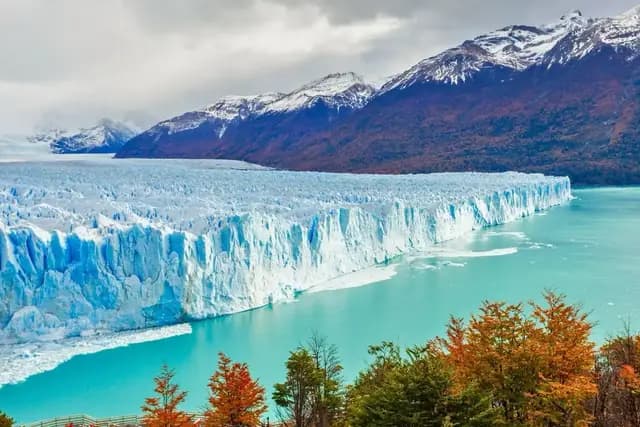 A vast, bright blue glacier face meets a turquoise lake, with snow-capped mountains and autumn foliage in the background under a cloudy sky.