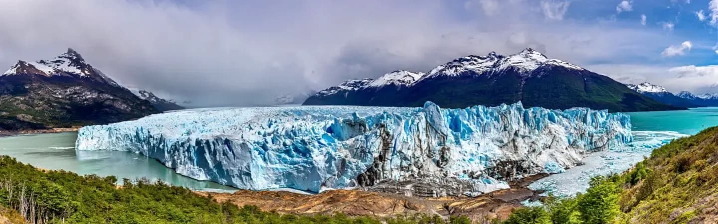 A vast, blue-tinged glacier juts into a turquoise lake, backed by snow-capped mountains and lush green hills under a cloudy sky.
