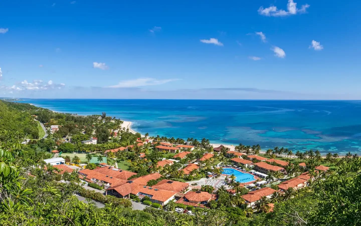 A high-angle view of a tropical resort with red-tiled roofs and a large blue swimming pool, nestled between lush green hills and a pristine white-sand beach meeting the turquoise ocean under a bright blue sky.